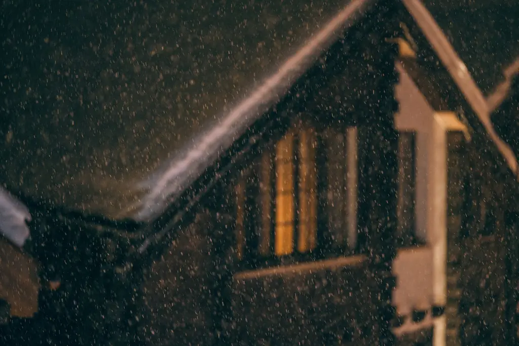 Snow falling on a house at night, with warm light in the windows